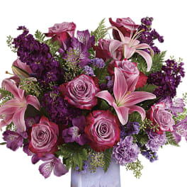 Pink lilies and roses with purple blooms arranged in a lavender glass vase