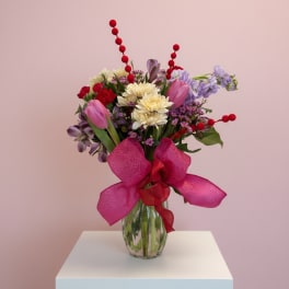 Mixed bouquet of pink, purple, and cream flowers in a glass vase with a large pink bow