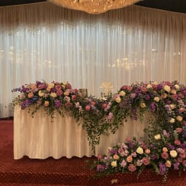 Low cascading arrangement of pink, purple, and white flowers decorating a long white table under a large chandelier