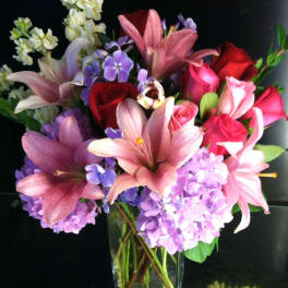 Pink lilies, red and pink roses, and purple hydrangeas in a clear glass vase