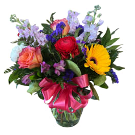 Bright mixed flower arrangement with yellow gerbera, roses, and red bloom in a clear vase with pink bow