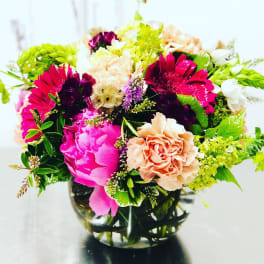 Bright mixed bouquet in a clear glass vase with pink, peach, white, and green blooms.