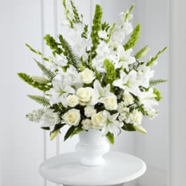 Tall white arrangement of lilies and roses with green accents in a white urn-style vase on a small round table