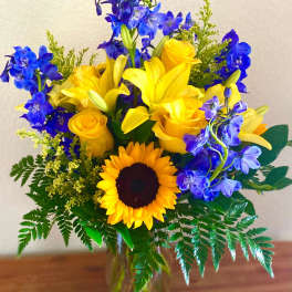 Yellow roses, lilies, and a sunflower in a glass vase with blue flowers
