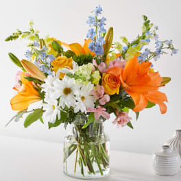 Bright mixed bouquet with orange lilies, yellow roses, and white daisies in a glass vase