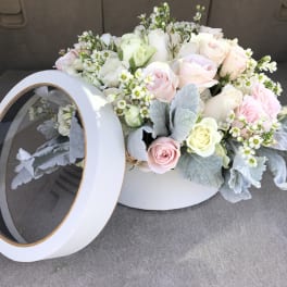 Pastel roses and small white flowers in a round white box with a clear lid.