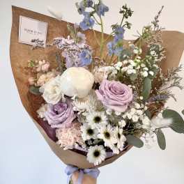 Pastel bouquet of lavender roses, white daisies, and mixed blooms wrapped in brown paper with a purple ribbon.