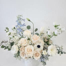 Low arrangement of ivory roses, white blooms, and pale blue flowers in a white ceramic vase