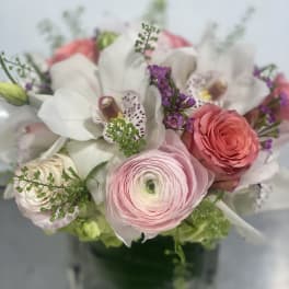 Short arrangement of pink ranunculus, roses, white orchids, and purple blooms in a clear glass cube vase