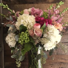 Tall glass vase of white hydrangeas, pink roses, orchids, and mixed blooms in soft and deep tones