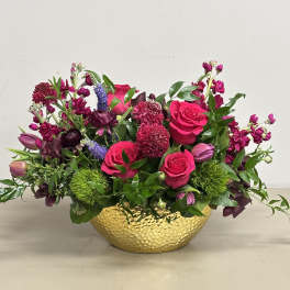 Low arrangement of hot pink roses and mixed purple flowers in a hammered gold bowl.