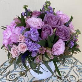Round arrangement of pink and lavender roses with mixed blooms in a white vase.