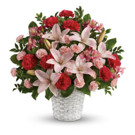 Pink lilies and red and light pink carnations arranged in a white woven basket.
