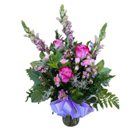 Mixed bouquet of pink roses, carnations, and tall snapdragons in a glass vase with a lavender ribbon bow.