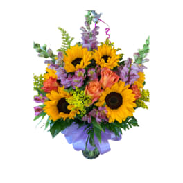 Mixed bouquet of sunflowers, orange roses, and purple flowers in a glass vase with a lavender bow