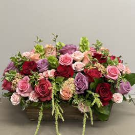 Low wooden box filled with red, pink, peach, and lavender roses with green accents.