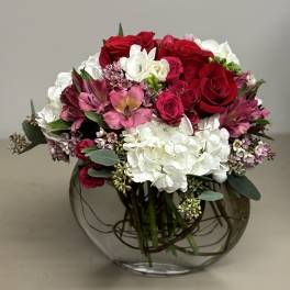 Low round arrangement of red roses, pink blooms, and white hydrangeas in a clear glass bowl vase