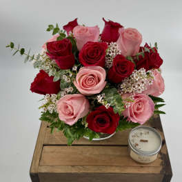 Low arrangement of red and pink roses in a glass vase on a wooden crate beside a small candle.