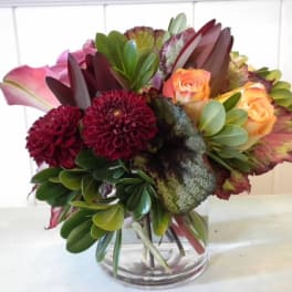 Compact arrangement of burgundy dahlias, peach roses, pink calla lilies, and variegated foliage in a glass vase