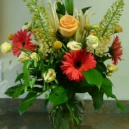 Mixed arrangement with red gerbera daisies, a peach rose, and cream flowers in a clear glass vase