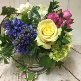 Short glass vase arrangement with yellow rose, pink roses, blue hyacinth, and white blooms