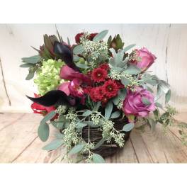 Low basket arrangement with pink roses, red mums, dark calla lilies, green hydrangea, and cascading eucalyptus.