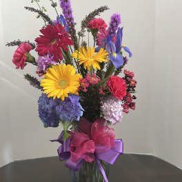 Tall mixed arrangement with yellow gerbera, blue hydrangeas, pink carnations and purple blooms in a glass vase.