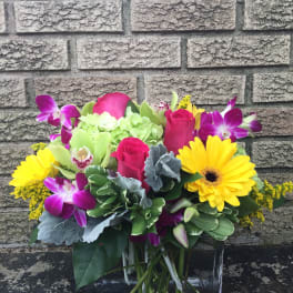 Bright mixed bouquet with yellow daisies, pink roses, and purple orchids in a glass vase