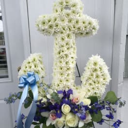 White floral cross on an easel with blue ribbon and purple flowers at the base