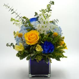 Bouquet of yellow and blue roses with white flowers in a square glass vase