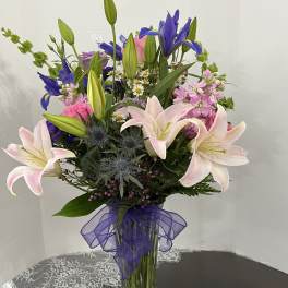 Mixed bouquet of lilies, irises, and pink flowers in a glass vase with a purple ribbon