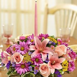 Low floral centerpiece with pink lilies, roses, and purple daisies around a tall pink candle.