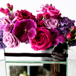 Pink and purple floral arrangement in a glass box with a dark ribbon