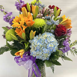 Mixed bouquet in a glass vase with blue hydrangea, roses, lilies, and purple flowers