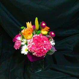 Bright mixed bouquet with pink hydrangea, orange roses, lilies, and daisies in a clear cube vase with purple wrap