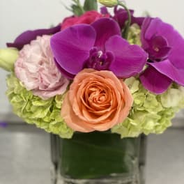 Low cube vase arrangement with purple orchids, peach rose, pink blooms, and green hydrangeas