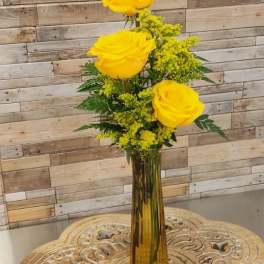 Tall amber vase with three yellow roses and yellow filler flowers on a carved wooden table.