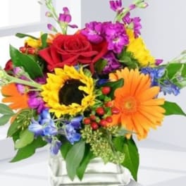 Colorful mixed bouquet in a clear square vase with a red rose, sunflower, and gerbera daisies