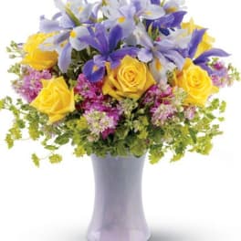 Mixed bouquet of yellow roses, purple irises, and pink flowers in a tall pale lavender vase