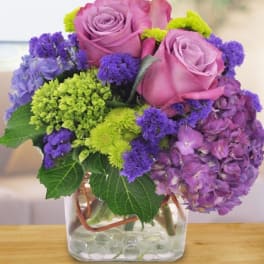 Low arrangement of purple hydrangeas, lavender roses, and green mums in a clear glass cube vase