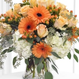 Arrangement of orange gerbera daisies, peach roses, and white hydrangeas in a clear glass vase