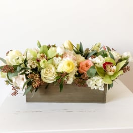 Low floral arrangement in a rectangular wooden box with cream, peach, and green blooms
