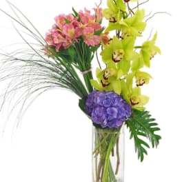 Tall arrangement of green orchids, pink alstroemeria, and purple hydrangea in a clear glass vase