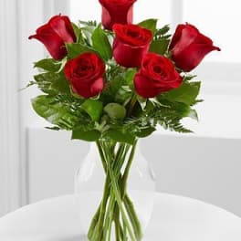 Six red roses in a clear glass vase on a white table.