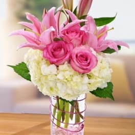 Pink lilies and roses with white hydrangeas arranged in a clear glass vase with pink accents