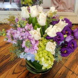 Low round arrangement of white tulips, green hydrangea, and purple lisianthus in a glass vase