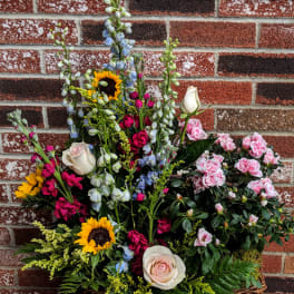 Tall mixed arrangement with sunflowers, roses, blue delphinium, and pink flowering plant in a wicker basket.