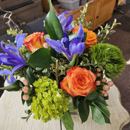 Low arrangement of orange roses, purple irises, and bright green blooms in a clear glass vase