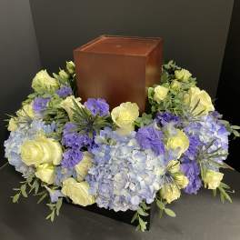 Floral wreath around a wooden urn with pale yellow roses and blue-purple hydrangeas