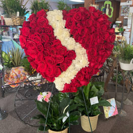 Tall heart-shaped standing spray of red roses with a white floral stripe above two potted green plants